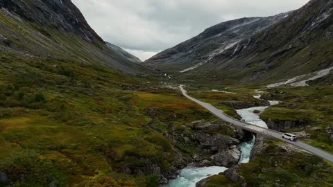 White VW camper van driving through dramatic mountain valley beside river in Vídeos de archivo 331978539