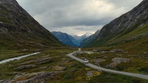 White VW camper van driving through dramatic mountain valley beside river in Vídeos de archivo 332215119