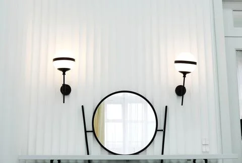 On a white wall hangs a round mirror in a black frame. Two black lamps hang.. Stock Photos