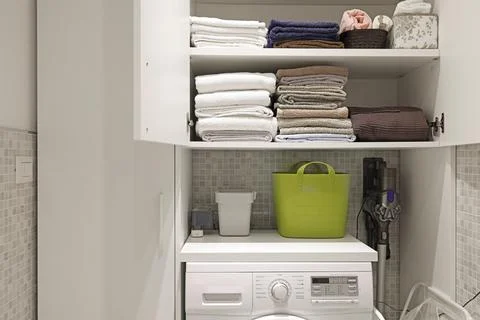 White washing machine below open cupboard shelves with neatly folded towels. Stock Photos
