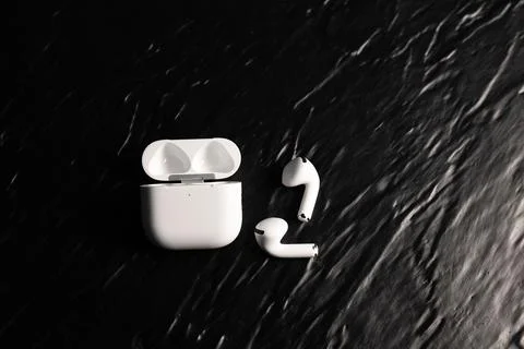 White wireless earbuds lying next to an open charging case on a black textu.. Stock-Fotos