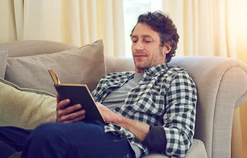 Who doesnt enjoy a good plot twist. a man reading a book on the couch. Fotos de archivo