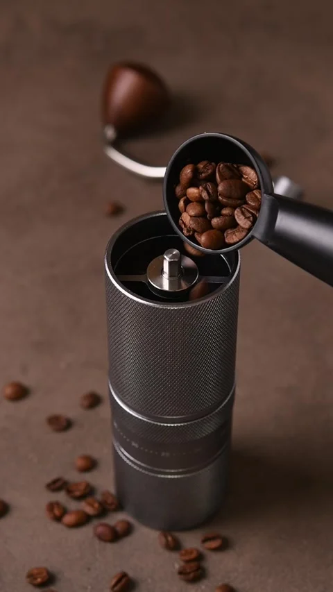 Whole coffee beans are slowly poured from a black scoop into a manual grinder on Stock Footage 346743277