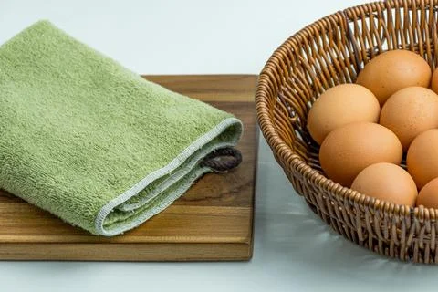 Wicker basket full of brown eggs next to folded kitchen towel isolated on whi Stock Photos