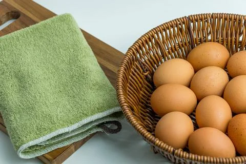 Wicker basket full of brown eggs next to folded kitchen towel isolated on whi Stock Photos
