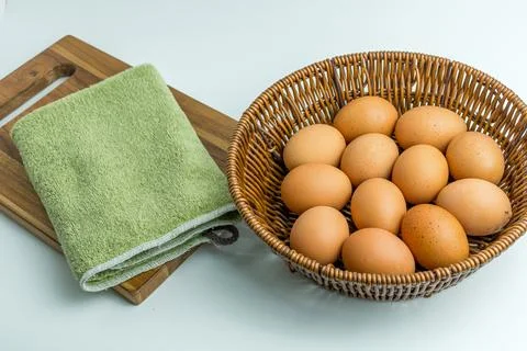 Wicker basket full of brown eggs next to folded kitchen towel isolated on whi Stock Photos