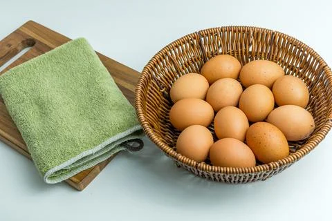 Wicker basket full of brown eggs next to folded kitchen towel isolated on whi Stock Photos