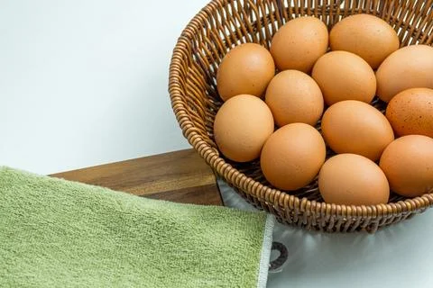 Wicker basket full of brown eggs next to folded kitchen towel isolated on whi Stock Photos