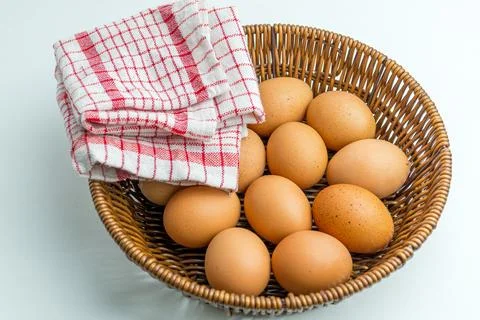 Wicker basket full of brown eggs next to folded kitchen towel isolated on whi Stock Photos