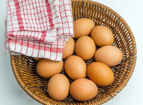 Wicker basket full of brown eggs next to folded kitchen towel isolated on whi Stock Photos