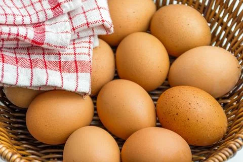 Wicker basket full of brown eggs next to folded kitchen towel isolated on whi Stock Photos