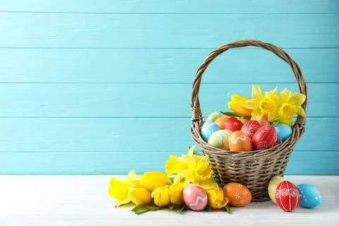 Wicker basket with painted Easter eggs and flowers on table against color bac Stock Photos