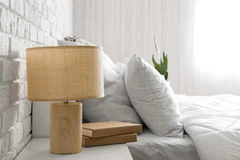 Wicker lamp with books on bedside table near bed against white brick wall in  Stock Photos