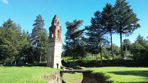Wide aerial orbit of a stone bell tower amid garden ruins and pines Stock Footage 346015666