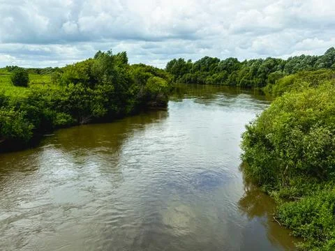 A wide, calm river with the reflection of clouds flows between green banks Stock Photos