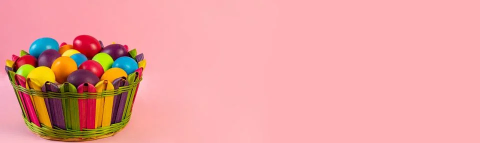Wide Easter banner. Multi-colored eggs lie in a wicker basket, pink background. Stock Photos