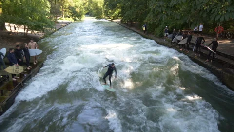 Wide of man riding Eisbach wave as other surfers wait on the side to jump in Vídeos de archivo 284498702