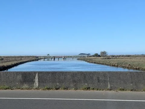 Wide river flowing through flat agricultural fields with distant rail bridge Stock Photos