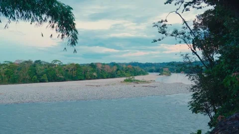 A wide river flows through lush Amazon rainforest with a rocky bank under a Video stock 345158113