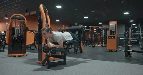Wide shot of 20s man training legs curls on machine in indoor fitness club Stock Footage 245696032
