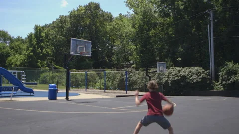 Wide shot of boy shooting hoops at outside baskeball court Stock Footage 133617821