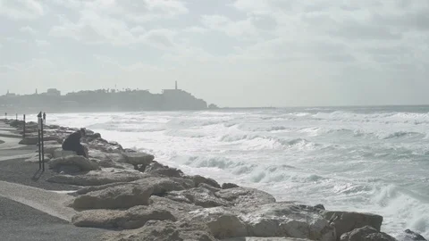Wide shot of an old man sitting on the rocks while waves crash Stock Footage 100705674
