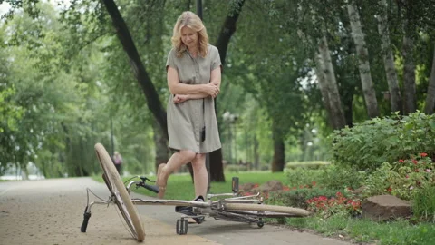 Wide shot of sad middle aged woman rubbing injured elbow and kicking bicycle Stock Footage 149082676