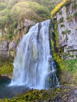 A wide spreading waterfall with many small steps flows from a rocky cliff Stock Photos