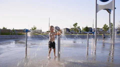 Wide view of 3 year old boy playing with yellow water gun at splash pad 스톡 동영상 233941036