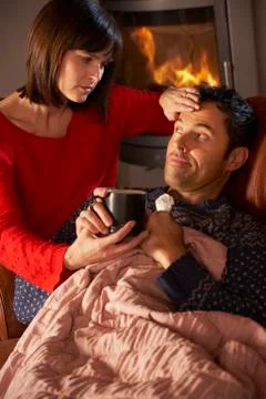Wife Nursing Sick Husband With Cold Resting On Sofa By Cosy Log Fire Stock Photos