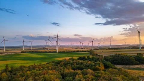 Wind farm beyond green fields and a belt of trees at dusk. White towers cro.. Stock Photos