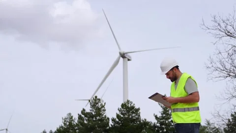 A wind farm technician checks notes on a clipboard, wearing a hard hat and sa Stock Footage 276312458