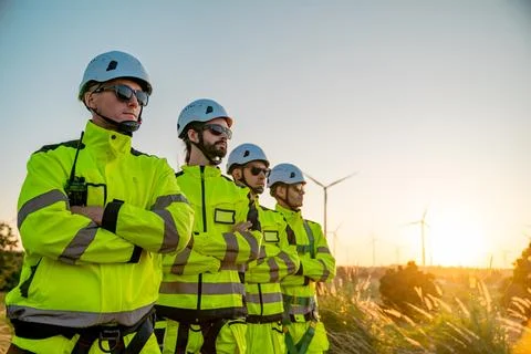 Wind farm technicians stand confidently during sunset wearing bright safety.. Stock Photos