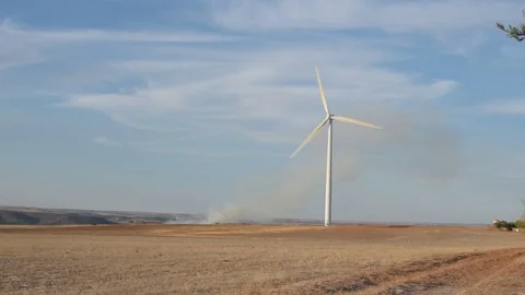 Wind turbine on open field with fire and smoke in the distance Stock Footage 80121059