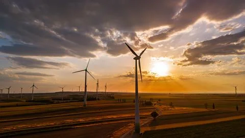 Wind turbine over fields beneath layered sunset clouds. The sun appears to .. Stock Photos