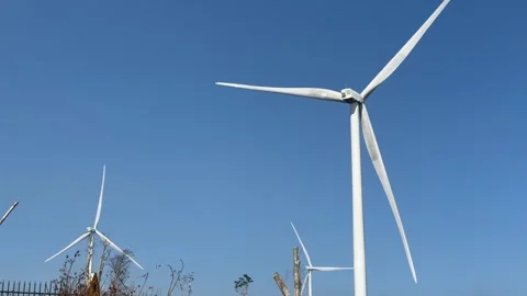 A wind turbine rotates on an elevated hilltop under a clear blue sky forming a 库存影片 333256365