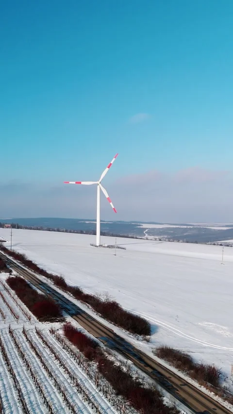Wind turbine rotating against a clear blue sky above a snow-covered field with a 스톡 동영상 340274105