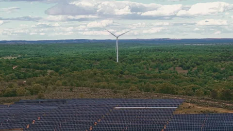 Wind turbine spinning over a forest with solar panel farm generating clean Видео 336213869
