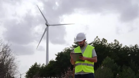 A wind turbine technician points at the wind turbine, holding a clipboard and Stock Footage 276312476