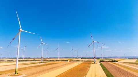 Wind turbines across parallel strips of harvested land. Red rotor tips rise.. Stock Photos