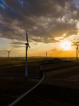 Wind turbines are seen on fields during sunset. The sky shows clouds with ora Stock Photos