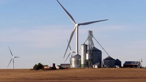 Wind turbines spin behind a large farm's grain storage facility Stock Footage 347405167