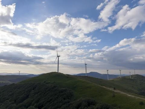 Wind turbines stand on a hill with clouds and blue sky at sunset Stock Photos