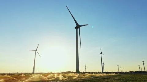 Wind turbines stand tall in a field while water sprays from irrigation systems Stock Footage 344265943