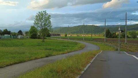 Winding asphalt path through green fields next to railway tracks under a cloudy Stock Footage 334865467