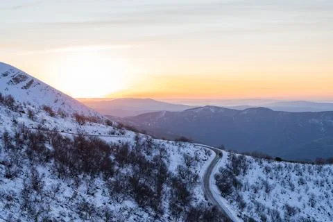 Winding mountain road passing through snowy landscape at sunrise Stock Photos