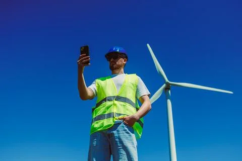 A windmill engineer in a reflective vest and hard hat stands near a wind turb Stock Photos