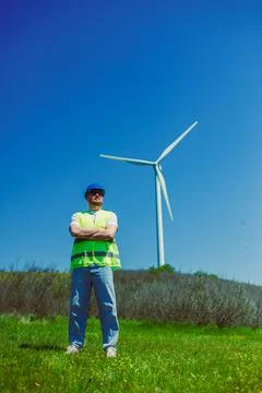 A windmill engineer in a safety vest stands with arms crossed in a grassy fie Stock Photos