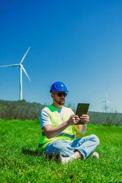 A windmill engineer sits on the grass in a field. He wears a safety vest and  Foto stock