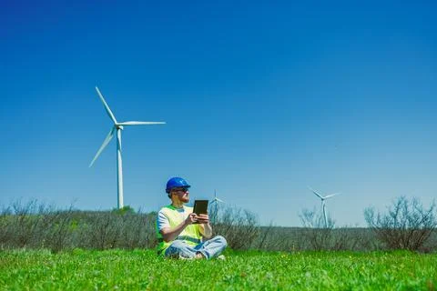 A windmill engineer sits on grass in a field while using a tablet. Wind turbi Stock Photos